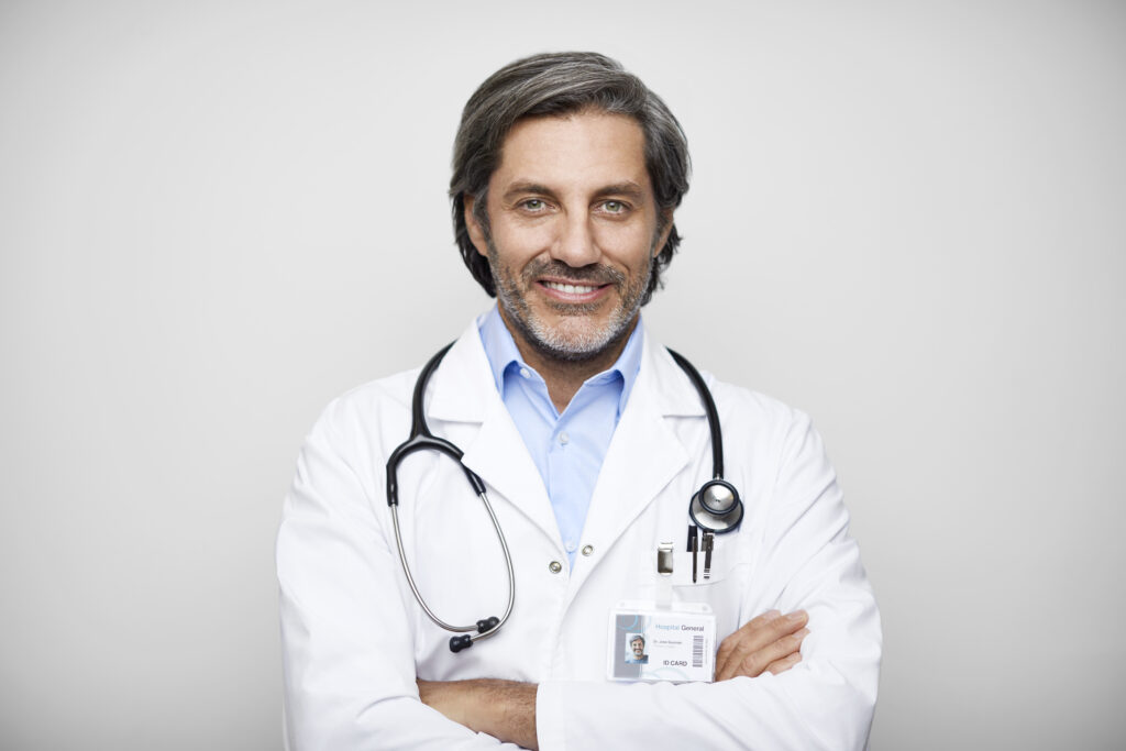Smiling mature Hispanic male doctor in lab coat. Portrait of smiling healthcare expertise is with arms crossed. He is against gray background.